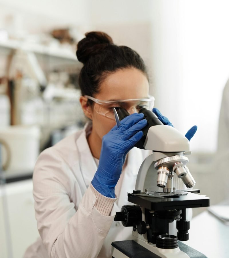 A female scientist in protective gear examines samples through a microscope in a laboratory setting.